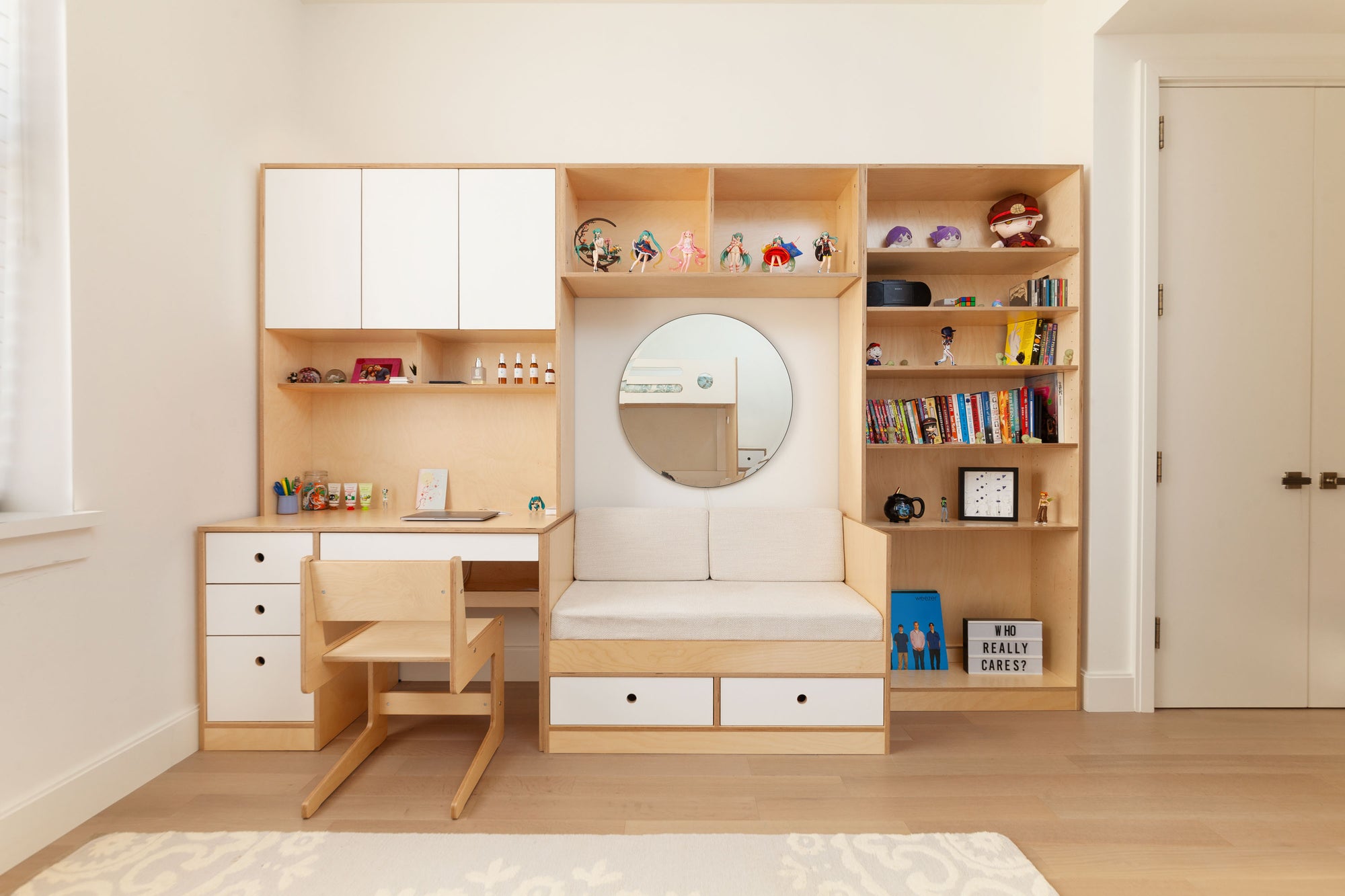 Integrated desk and seating unit with white oak finish and white painted accents. Features a compact desk area with drawers, upper cabinets, and open shelving. The seating section includes two drawers underneath and a large round mirror above, with additional shelves for books and decorative items.

