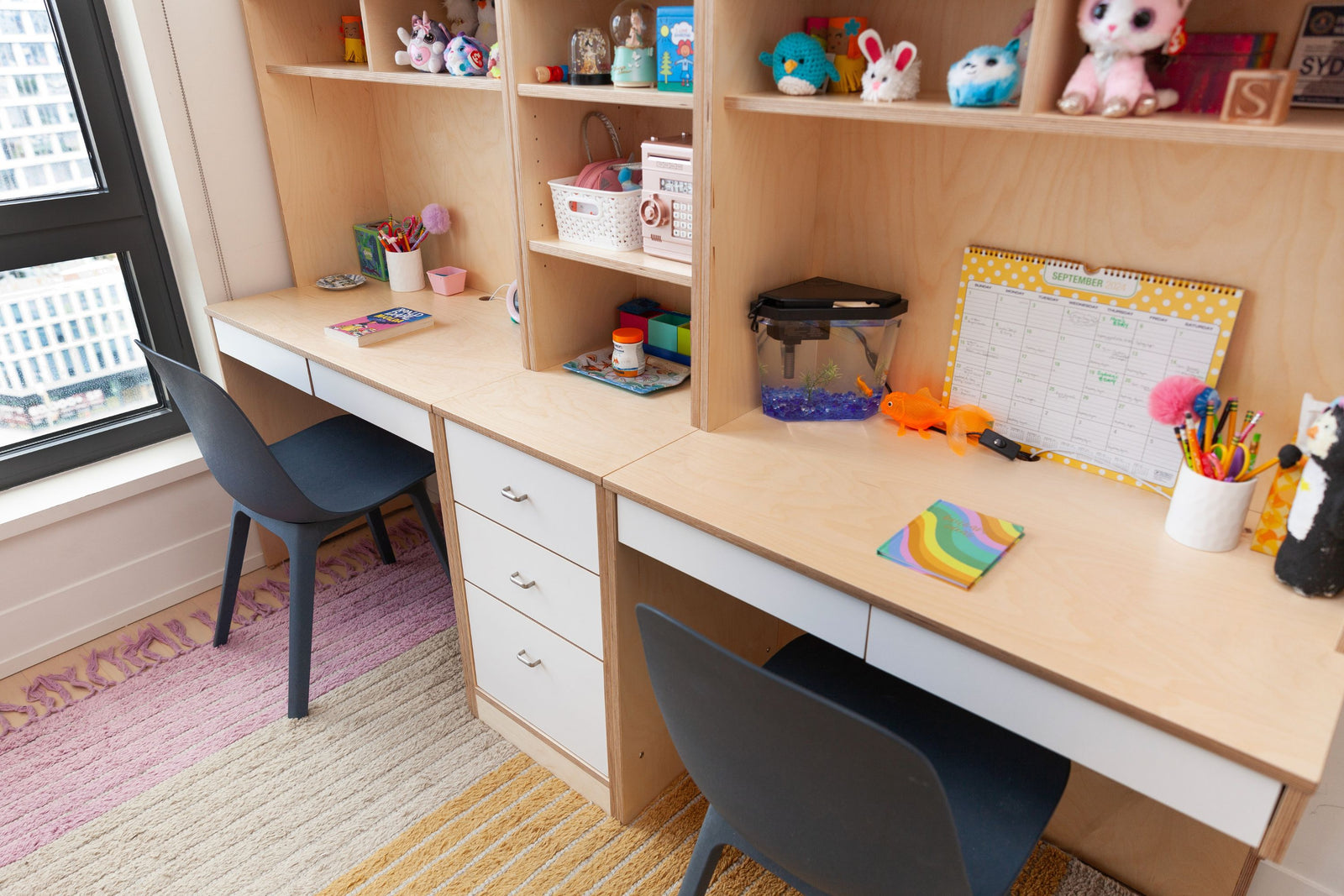 A pair of custom-built desks with a shared center storage unit and upper shelving, designed in a light birch finish. Each workspace includes drawers, open shelving, and personal decorations like plush toys, books, a fish tank, and stationery. The desks are complemented by modern dark blue chairs and colorful rugs, creating a cozy and organized study area.