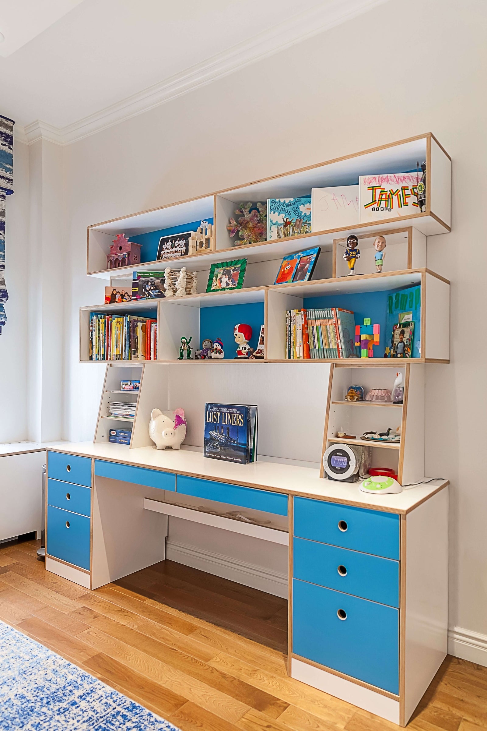A custom-built Dumbo desk featuring blue and white cabinetry, multiple drawers, and open shelving for storage. The desk has a spacious workspace with built-in shelves above, displaying books, toys, framed photos, and decorative pieces. The design combines functionality with playful elements, making it ideal for a child's study or creative space.