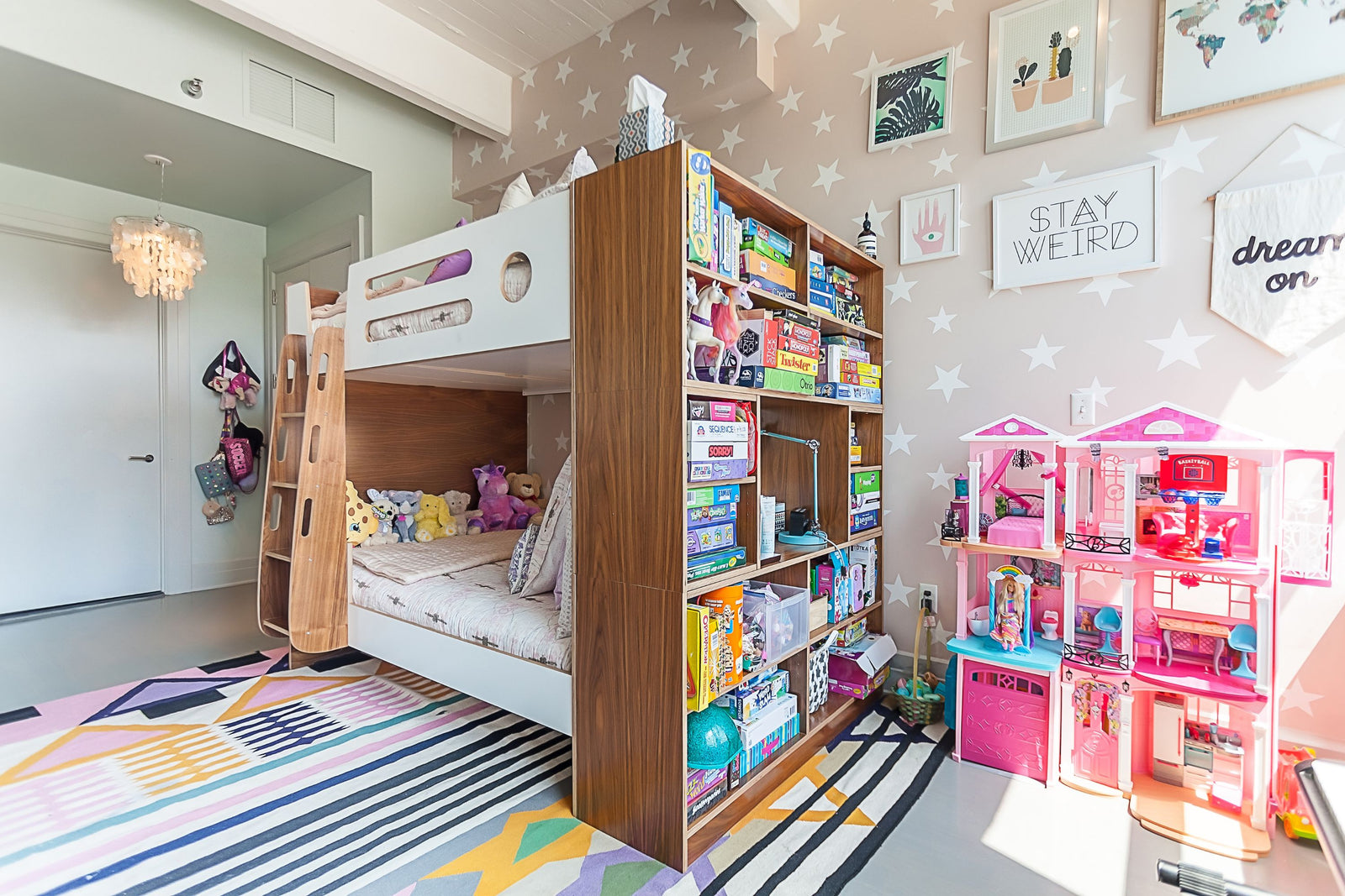 A stylish full-size bunk bed in walnut and white, featuring a built-in bookcase at the foot of the bed filled with board games, books, and toys. The room has a playful, modern aesthetic with star-patterned wallpaper, a colorful geometric rug, and a large pink dollhouse, creating a cozy and organized space for a child.