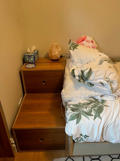 wooden nightstand with double drawers, beside a bed. A sloth plush toy is sitting on the nightstand