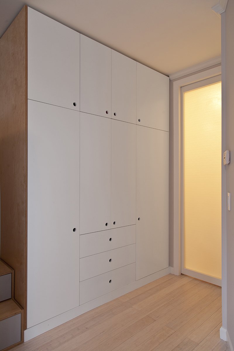 Sleek white built-in wardrobe in a minimalist corridor with wooden flooring and a softly lit room adjacent.