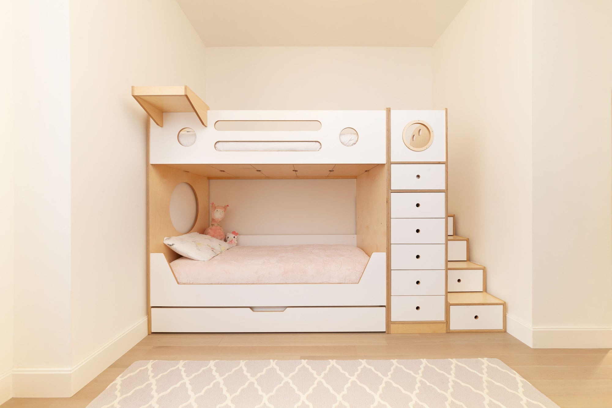 Minimalist kids' bunk bed in a birch and white finish, featuring built-in storage drawers and a staircase with integrated storage steps. The cozy lower bunk includes circular cutouts and a soft pink bedding set, creating a calm and functional sleeping area for children.

