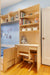 Children's room desk with built-in shelving, light wood finish, and adjacent bed. The shelves are stocked with books, toys, and storage bins, creating a functional study and play space.

