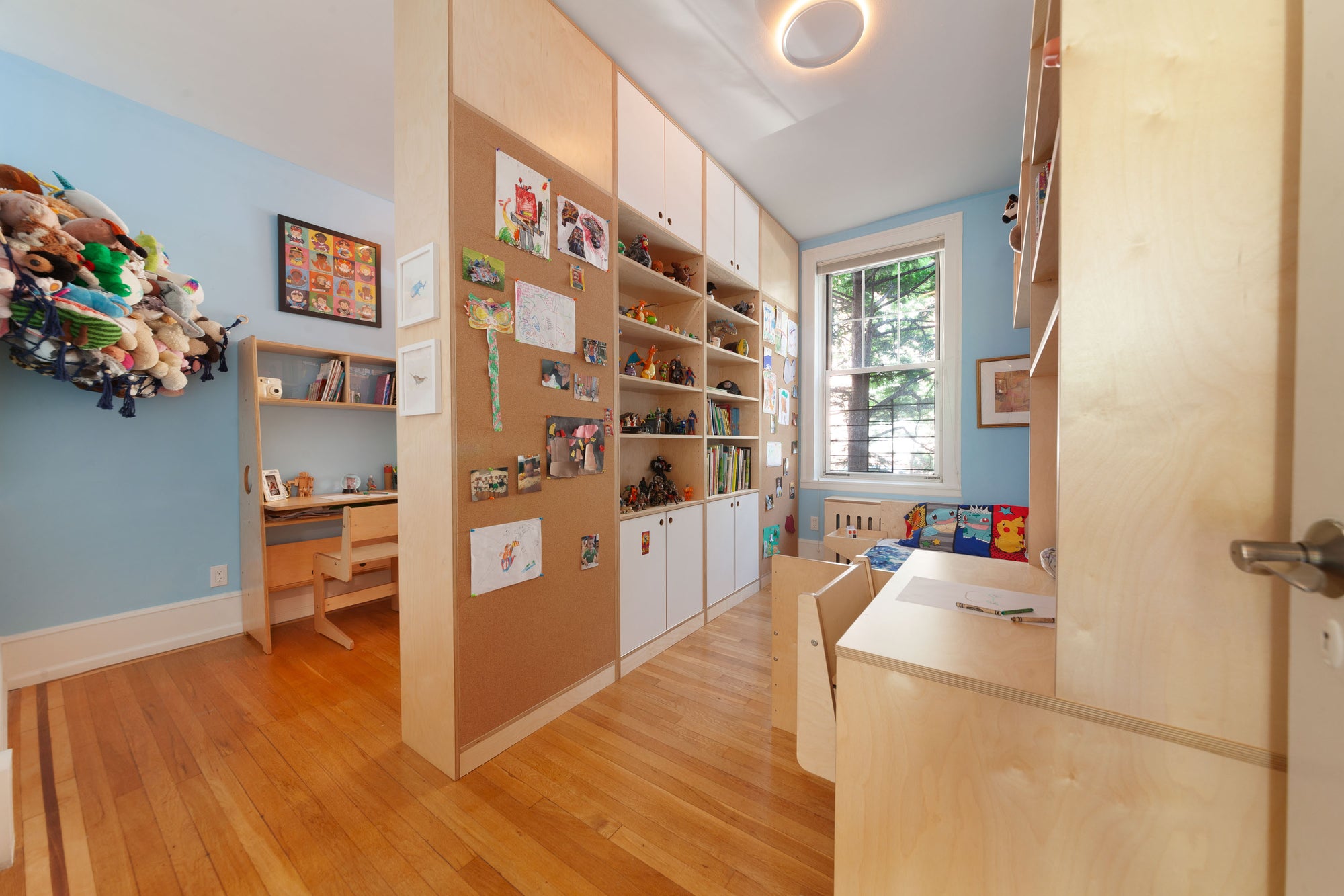 Children's room with birch shelving, a corkboard partition wall displaying artwork, and two desks in a blue and natural wood space.

