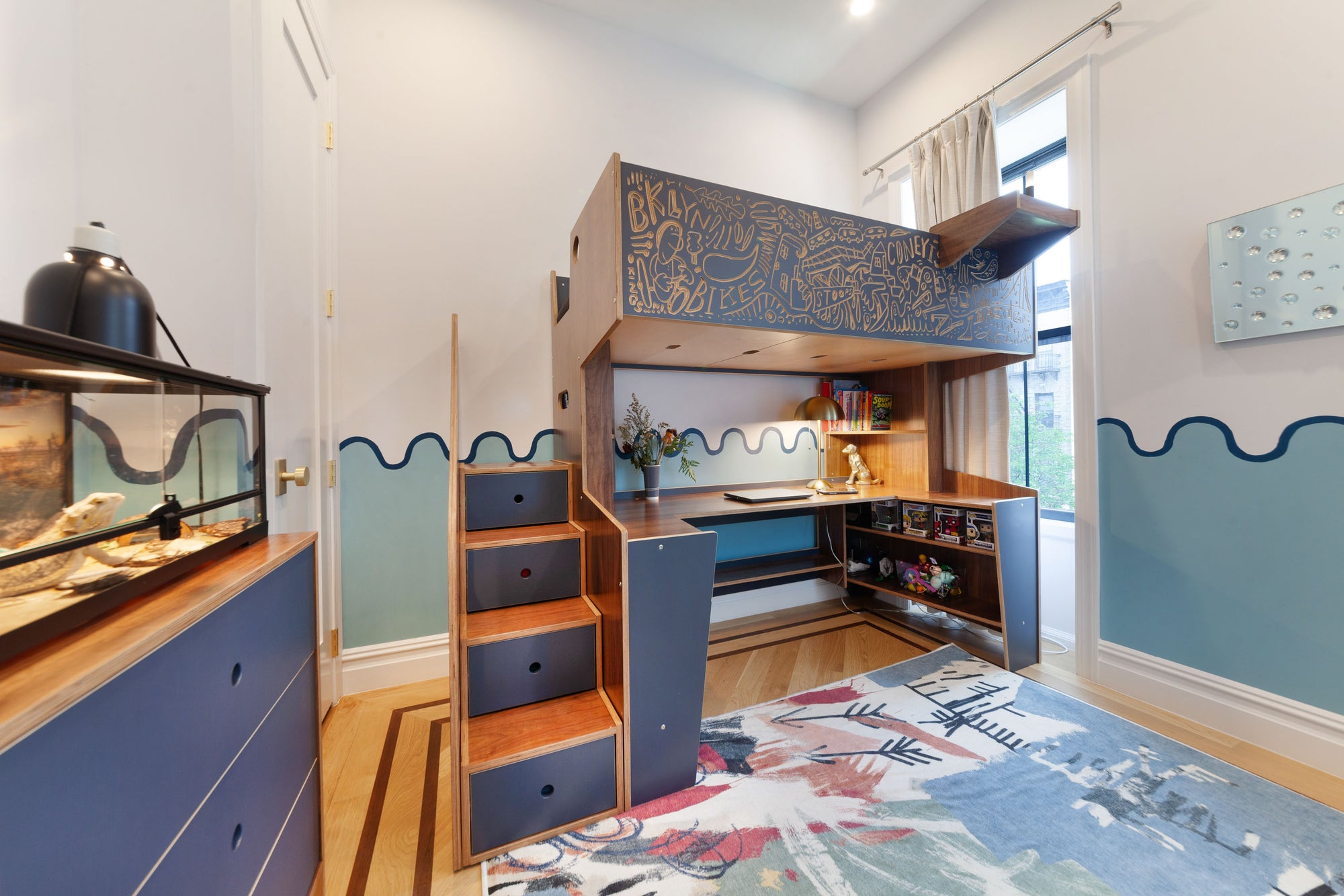 Custom loft bed with integrated desk and storage, featuring a unique artistic design on the loft exterior in blue and wood tones. The staircase includes built-in storage drawers, and the room is decorated with wave-inspired wall art, creating a stylish and functional workspace for kids.

