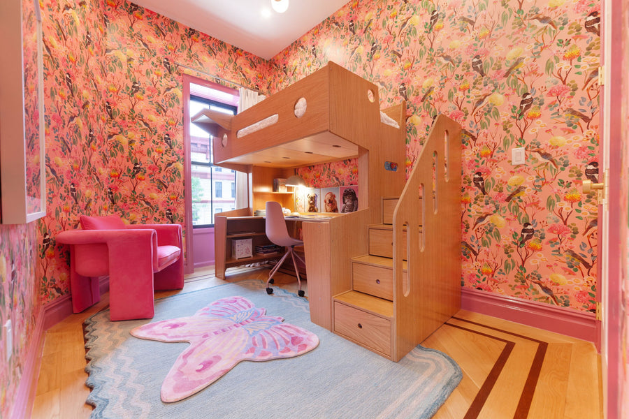 Children's room with a loft bed featuring an integrated desk and storage steps, set against vibrant pink floral wallpaper. The cozy workspace below includes books and decor, with a stylish pink accent chair and a butterfly rug, creating a lively and imaginative study and sleep area.
