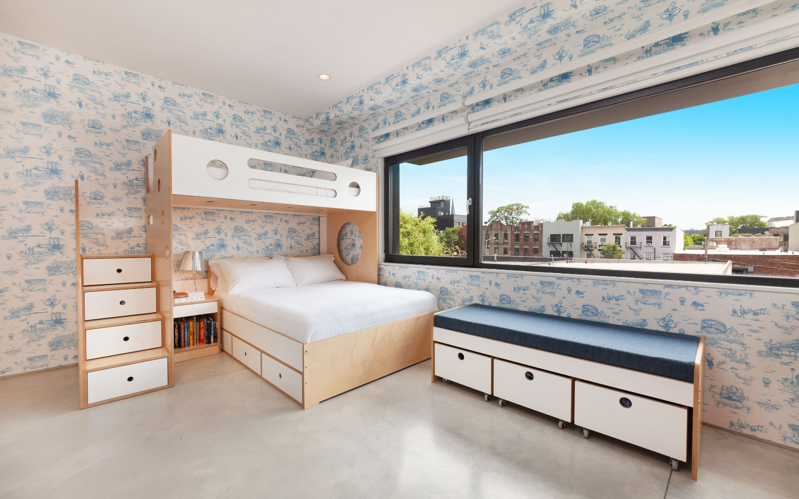 Bright children's room featuring the Marino L-shaped bunk bed in birch with white accents, built-in storage drawers under the bed, and staircase drawers. The room includes a window bench with blue cushions and additional storage drawers, set against playful blue-and-white wallpaper with city views, creating a functional and airy space.

