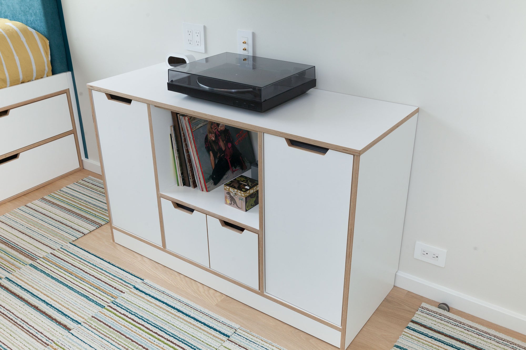 White media cabinet with built-in record storage and a turntable on top, next to a bed with storage drawers.

