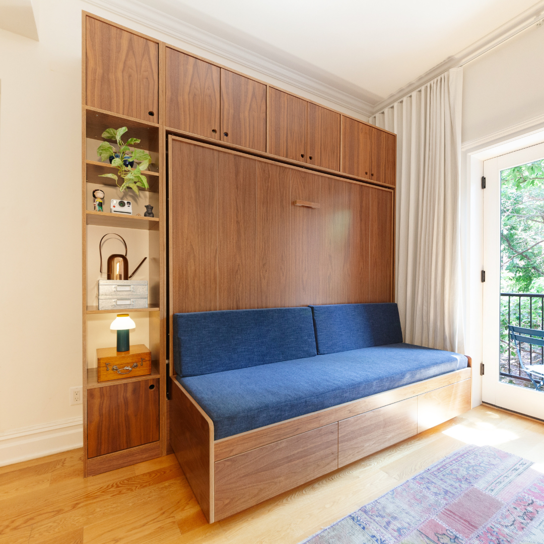 Murphy bed in closed position with walnut finish, featuring a blue sofa seat and adjacent open shelving for storage in a bright room with natural light.