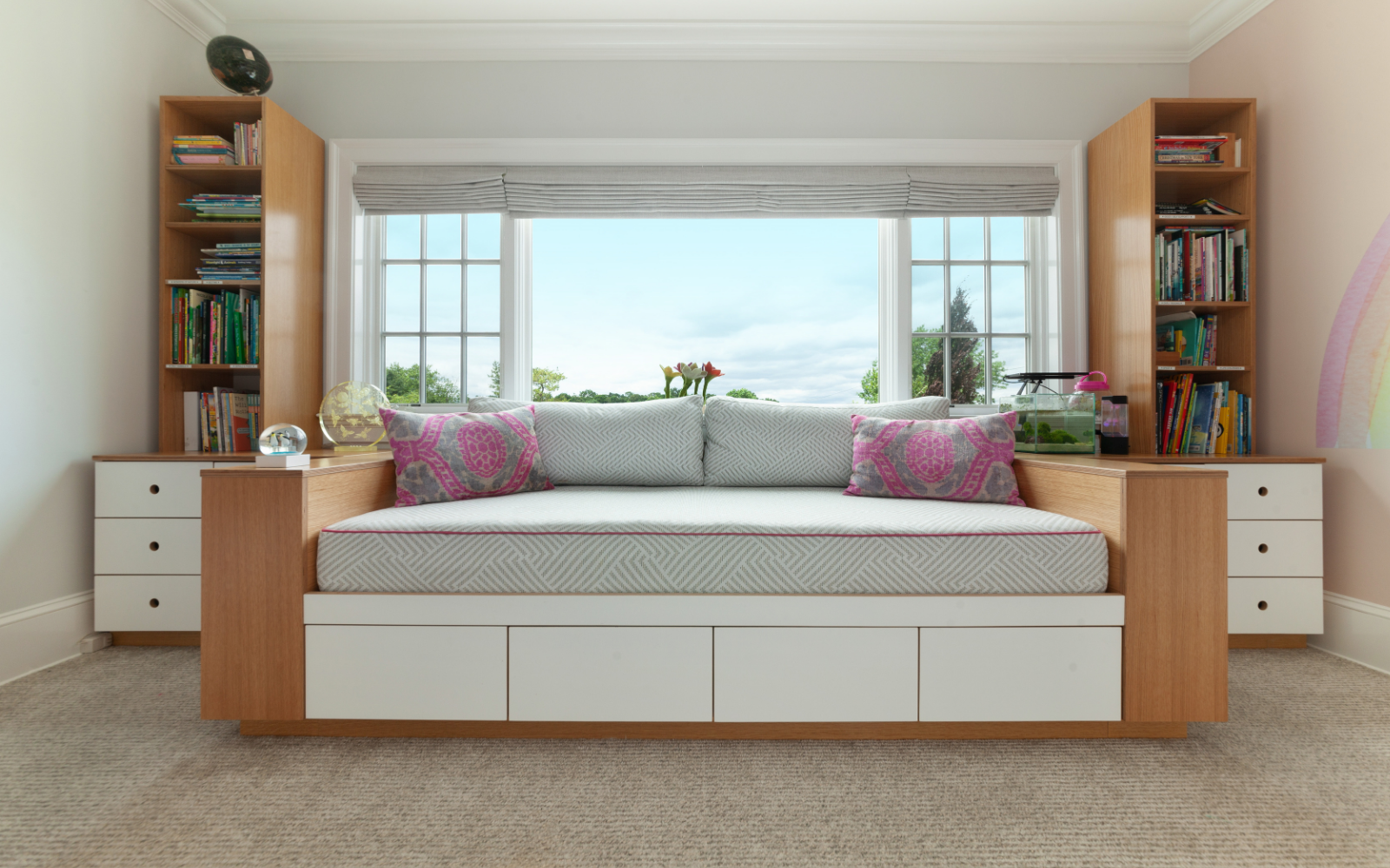 Custom oak and white bench with integrated storage in a cozy room. The bench is positioned under a large window, offering a relaxing spot with natural light and views of greenery. The design features a central seating area with a geometric-patterned cushion and accent pillows in pink and gray. Flanking the bench are open shelving units and cabinets in oak, holding books and small decorative items. Drawers beneath the bench provide additional storage, blending seamlessly into the white base. The room is soft