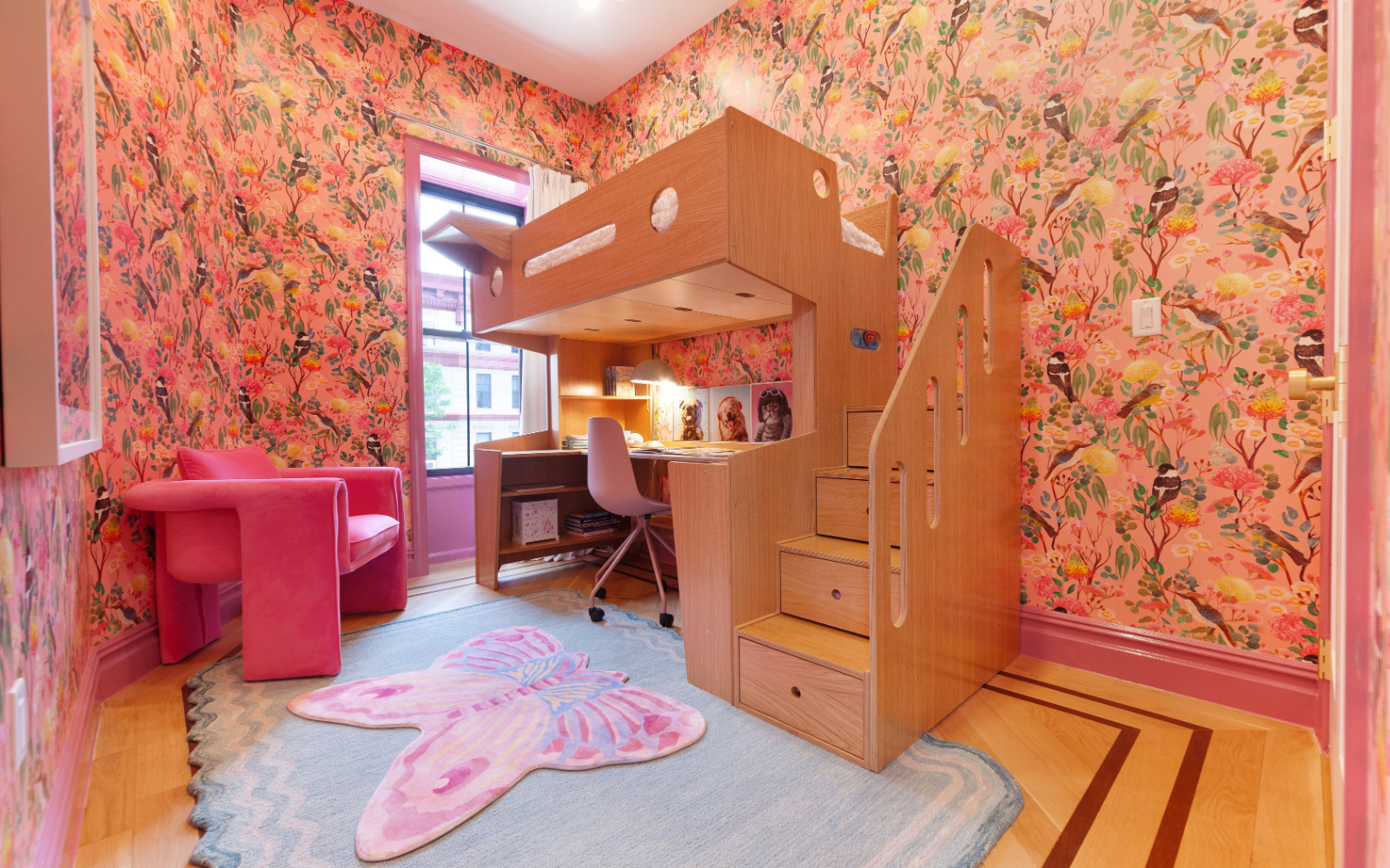 Custom oak loft bed with integrated desk and staircase storage set against vibrant floral wallpaper. The loft bed provides a cozy sleeping area above, with cut-out accents for a playful design. Beneath the bed, a spacious desk offers ample workspace, illuminated by a desk lamp. The staircase leading to the loft includes drawers for additional storage. A pink chair and butterfly-shaped rug complement the room's lively decor, adding charm and color to the space. The setup is designed for both functionality an