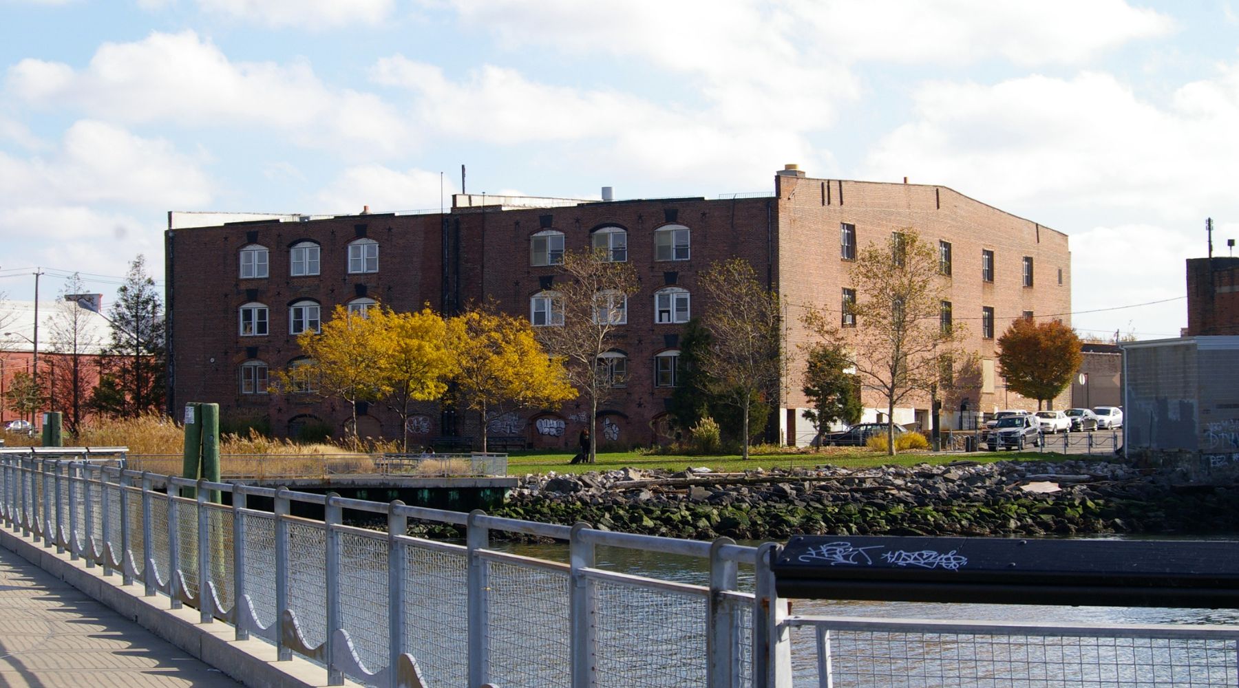 A scenic view of a historic brick warehouse in Red Hook, Brooklyn, captured from a waterfront walkway. The building features arched windows and graffiti on the lower walls, surrounded by trees with autumn foliage. The foreground includes a metal railing along the water’s edge, with a peaceful urban-industrial backdrop.