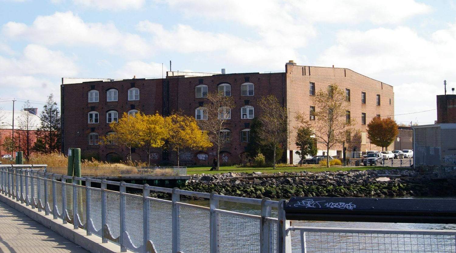 Urban scene with a Casa Kids warehouse along the waterfront and pier, trees, and a fence under a Blue sky with clouds.