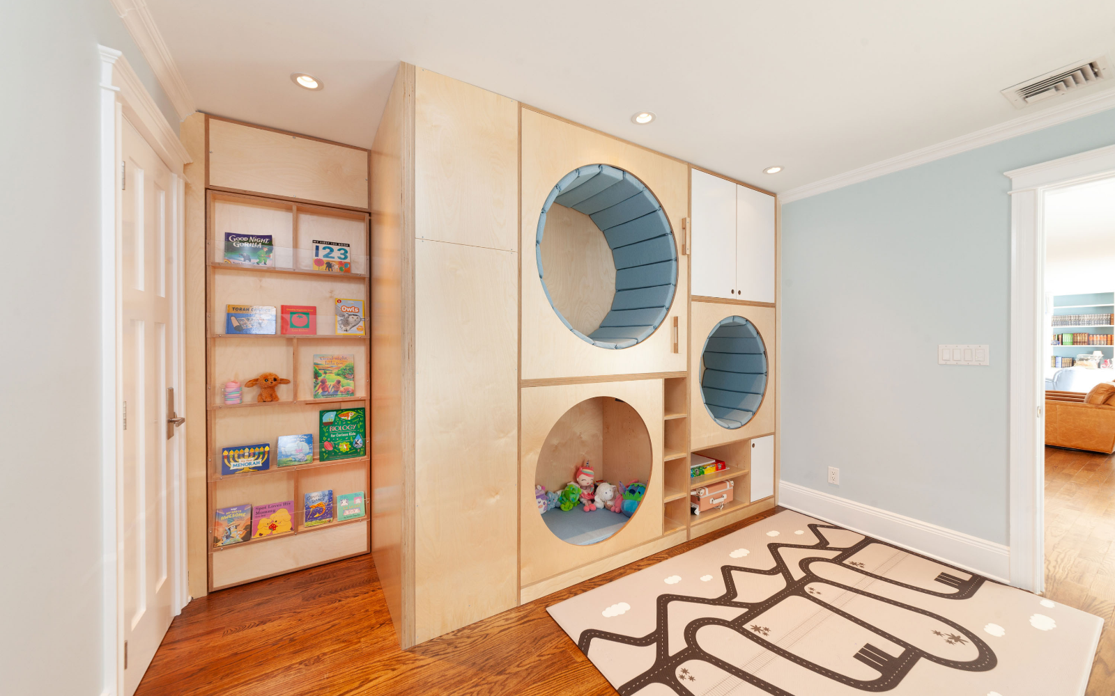 Custom room partition designed for a kids' play area, featuring birch wood with circular cubbies padded in blue for cozy seating or play spaces. Adjacent is a wall-mounted book display with open shelving for easy access to books and toys. The setup combines functionality and fun, creating a dedicated space for reading and imaginative play.

