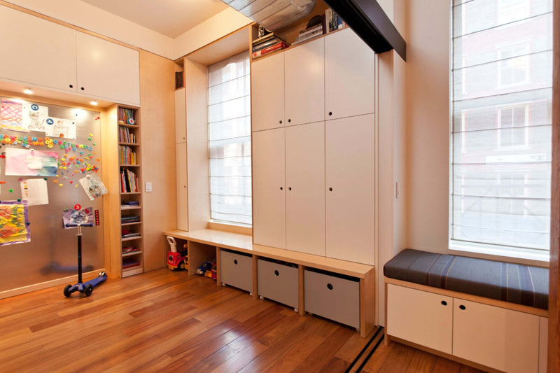 Custom-built cabinetry storage with a combination of upper cabinets, lower storage bins, and a built-in window bench with cushioned seating. The room features a sleek, modern design with birch and white finishes, maximizing functionality and organization in a bright and inviting space.