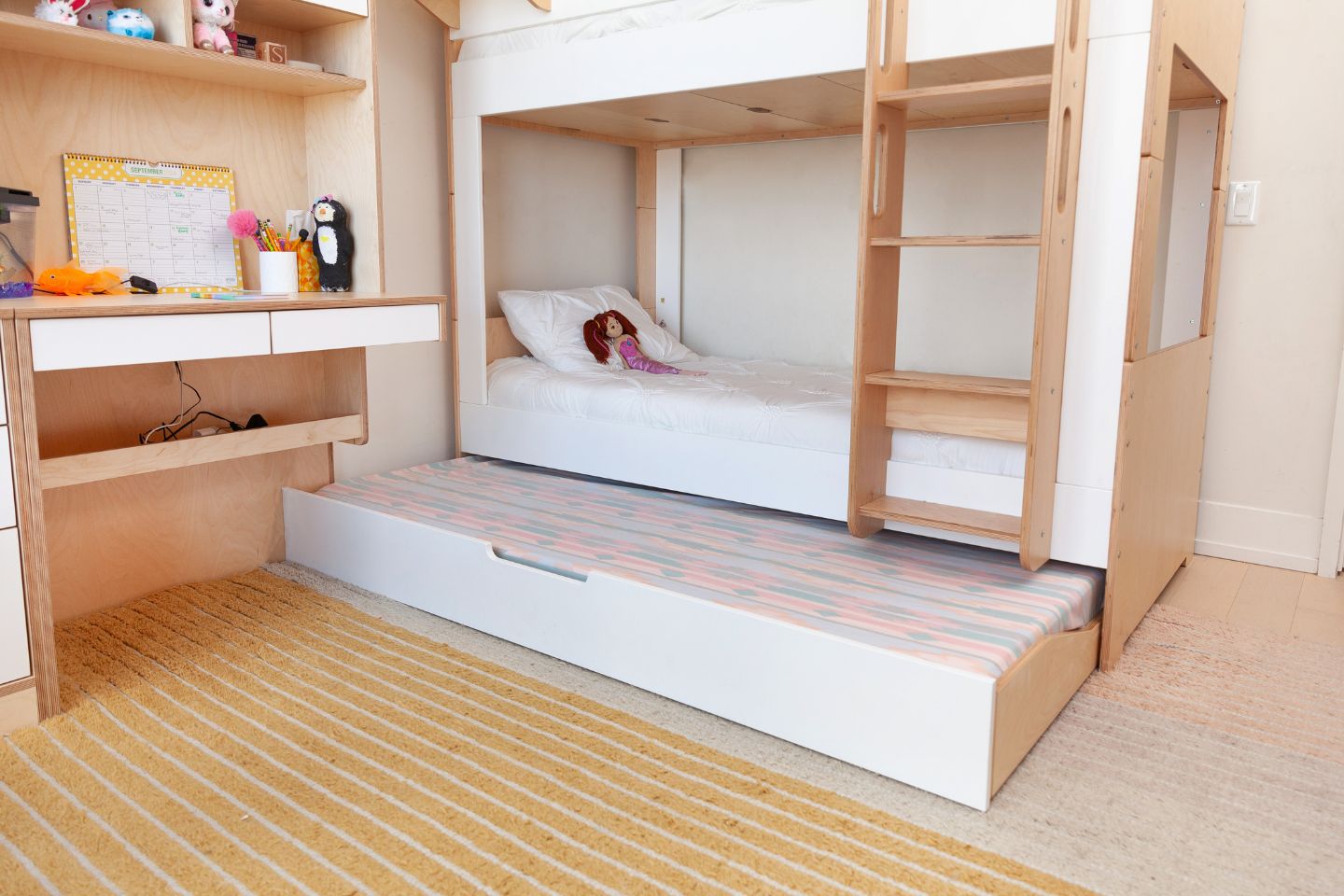 Space-saving trundle bed on wheels in Birch wood with white accents designed to tuck neatly under a bed frame on bottom of twin mattress with white wall and matching desk.