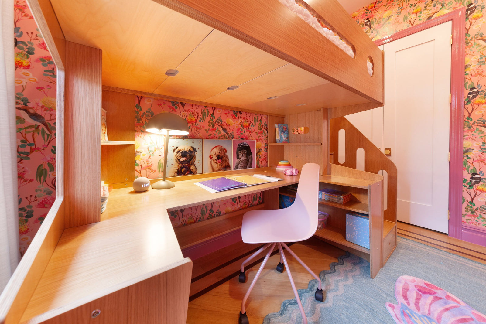 A cozy built-in desk beneath the Marino loft bed, featuring warm wood tones and integrated shelving. The workspace is decorated with colorful floral wallpaper, playful animal artwork, and a pink swivel chair. Soft lighting and neatly arranged books and storage boxes create a functional and inviting study area.