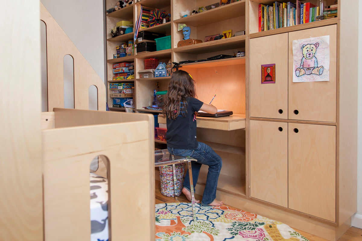 A child sits at a wooden desk in a cozy study area with built-in shelves filled with toys, books, and art supplies.
