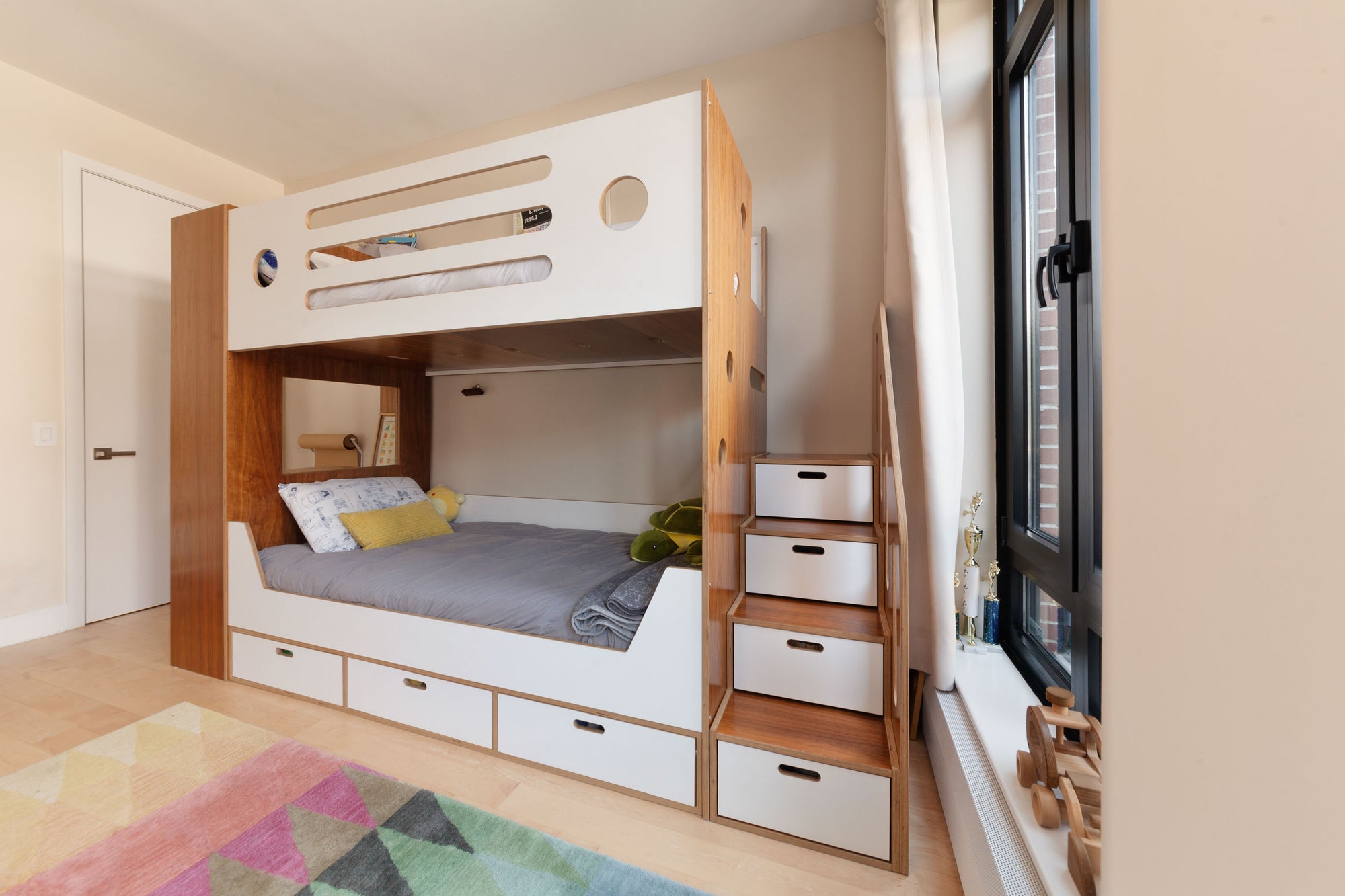 Custom bunk bed in a walnut finish with white accents, featuring integrated storage drawers under the lower bunk and in the staircase. The design includes circular cutouts on the upper bunk and a cozy lower bunk setup, providing a stylish and functional solution for a children’s bedroom.

