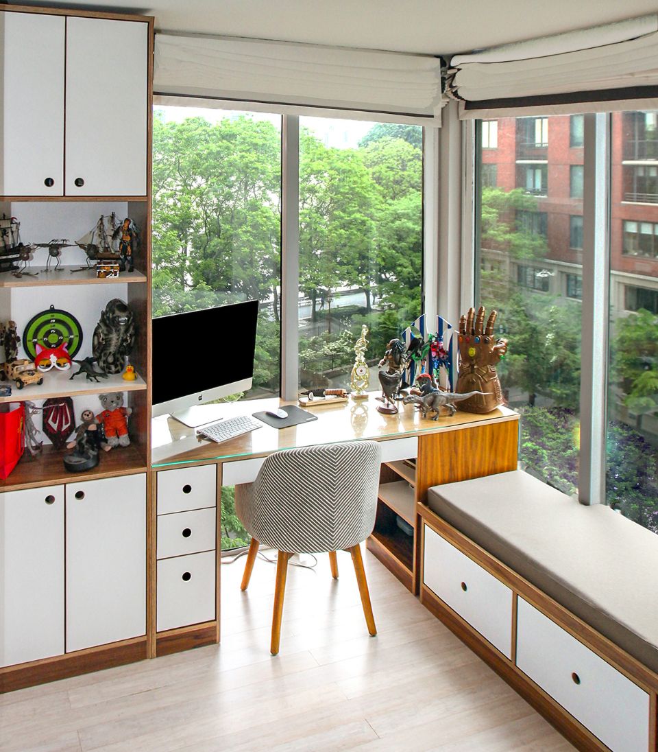 A modern workspace featuring a custom-built walnut desk with integrated storage drawers, open shelving, and a seating area with built-in storage. Large windows provide ample natural light and a view of lush greenery. The space is decorated with collectibles, action figures, and unique display pieces, blending functionality with personal style