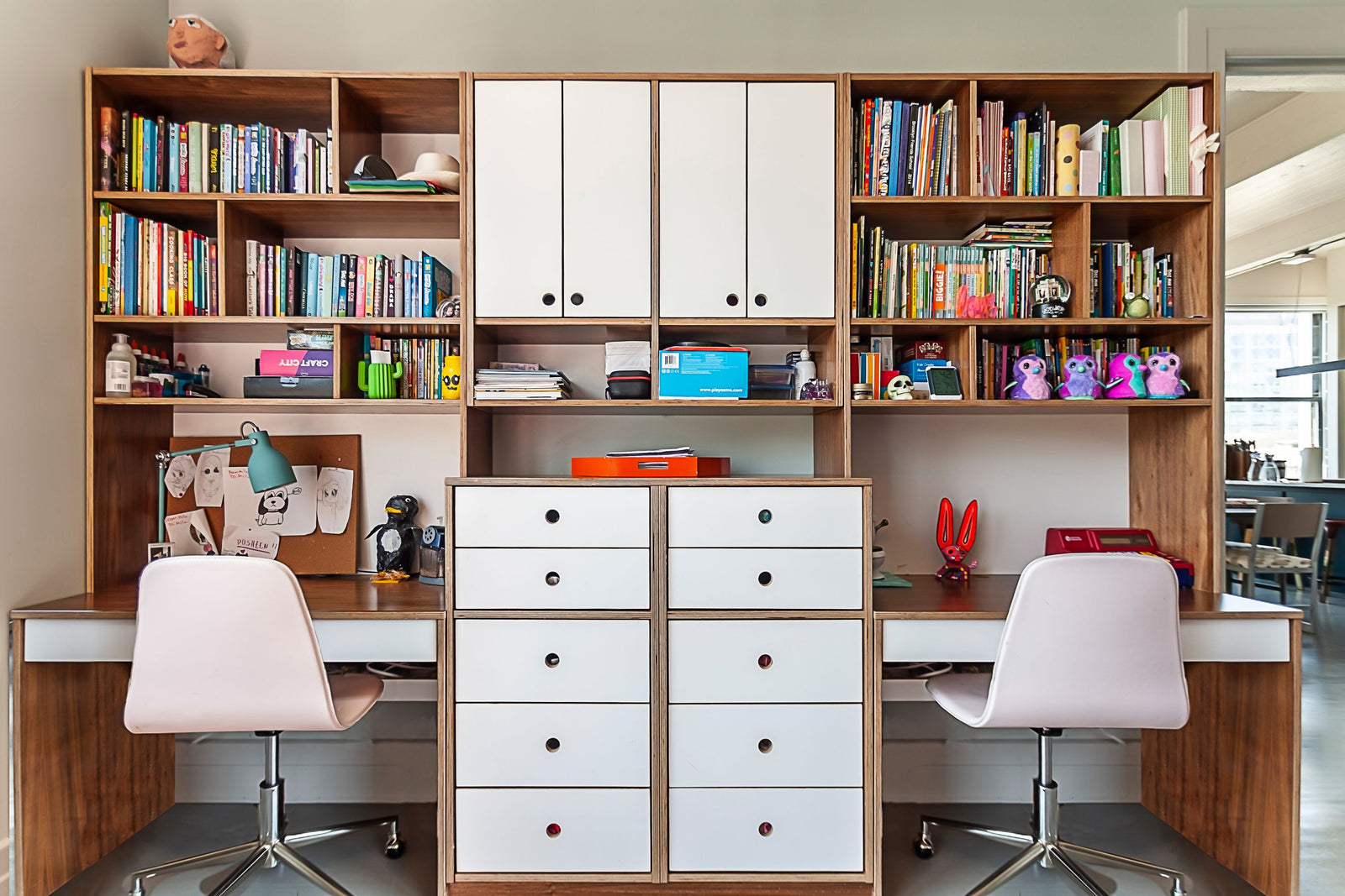 A custom-built shared workspace featuring walnut and white desks with a central dresser for storage. Each desk has open shelving for books, personal items, and decor, along with upper cabinets for additional organization. The design is both functional and stylish, with modern white chairs and a well-organized layout for studying or creative activities
