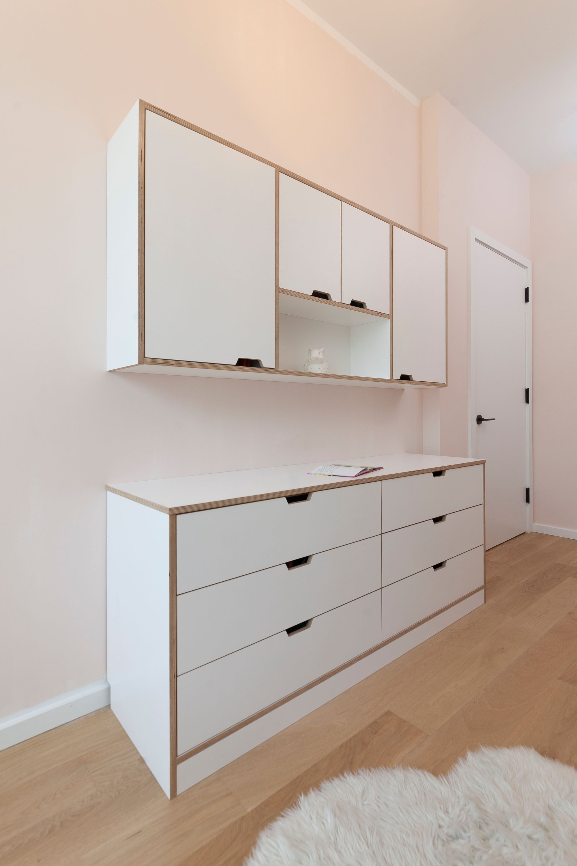 White dresser unit with built-in drawers and a wall-mounted hanging storage cabinet in a light pink room.

