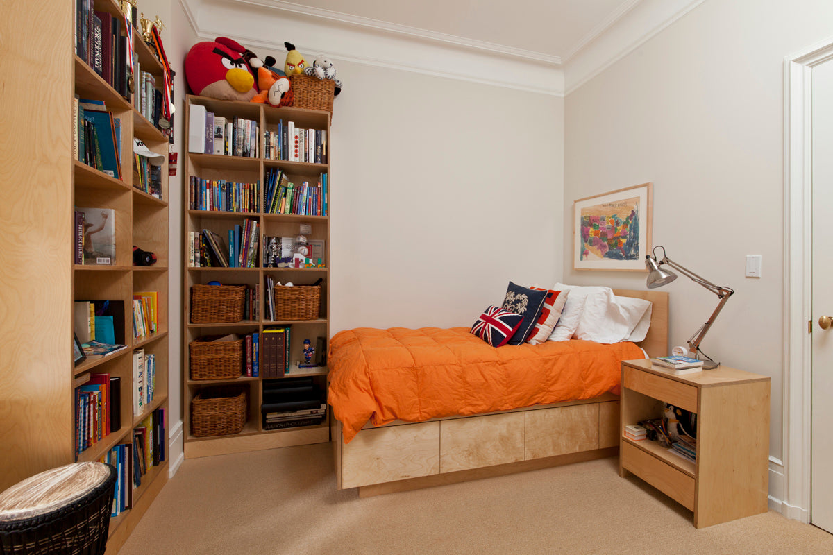 A cozy bedroom with a wooden bed, orange blanket, bookshelf filled with books and baskets, and a nightstand with a lamp.