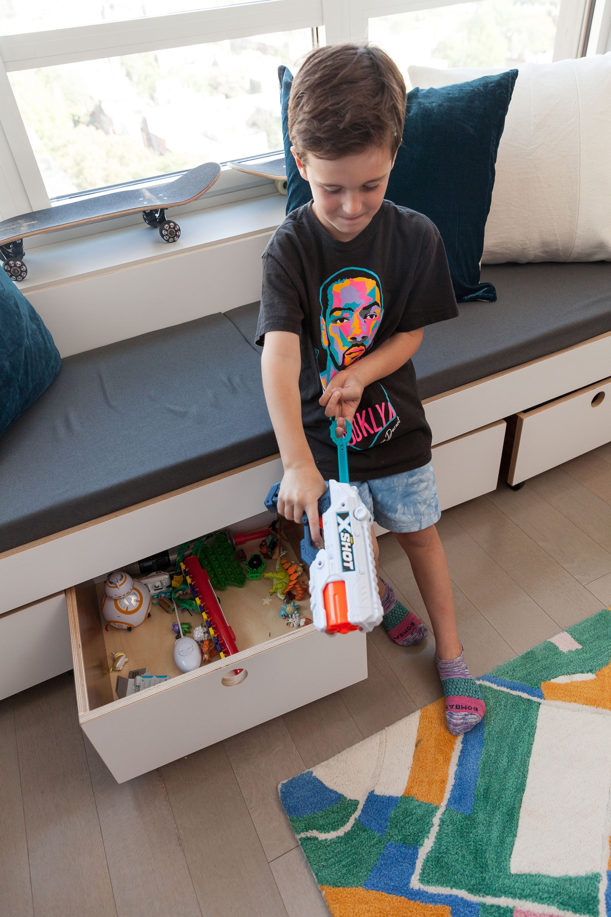 Child playing next to a window bench with pull-out storage drawers, filled with toys and action figures. The bench features a cushioned seat and is finished in white with birch accents, providing a practical and organized play area.

