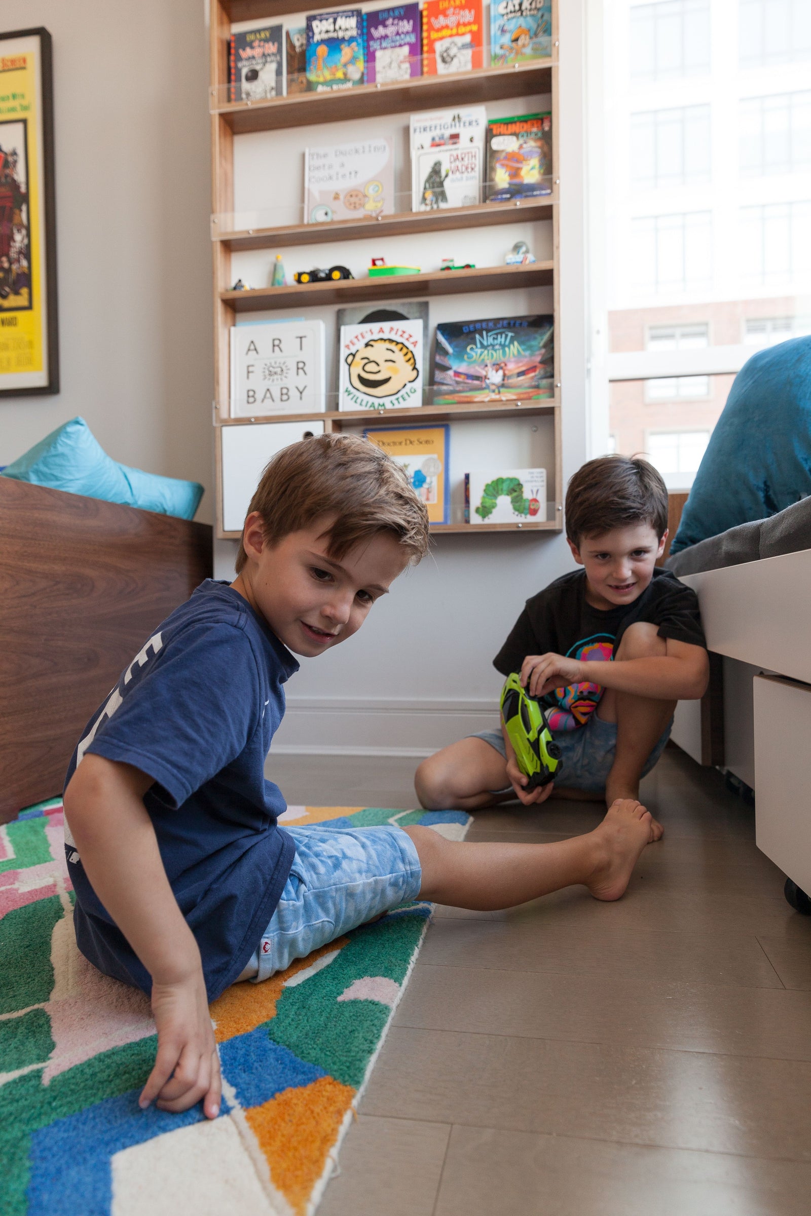 Two young boys play on a colorful geometric rug near a modern window seating area with built-in storage. A sleek hanging storage cabinet in walnut and white displays an array of children's books and toys, adding both function and charm to the space.