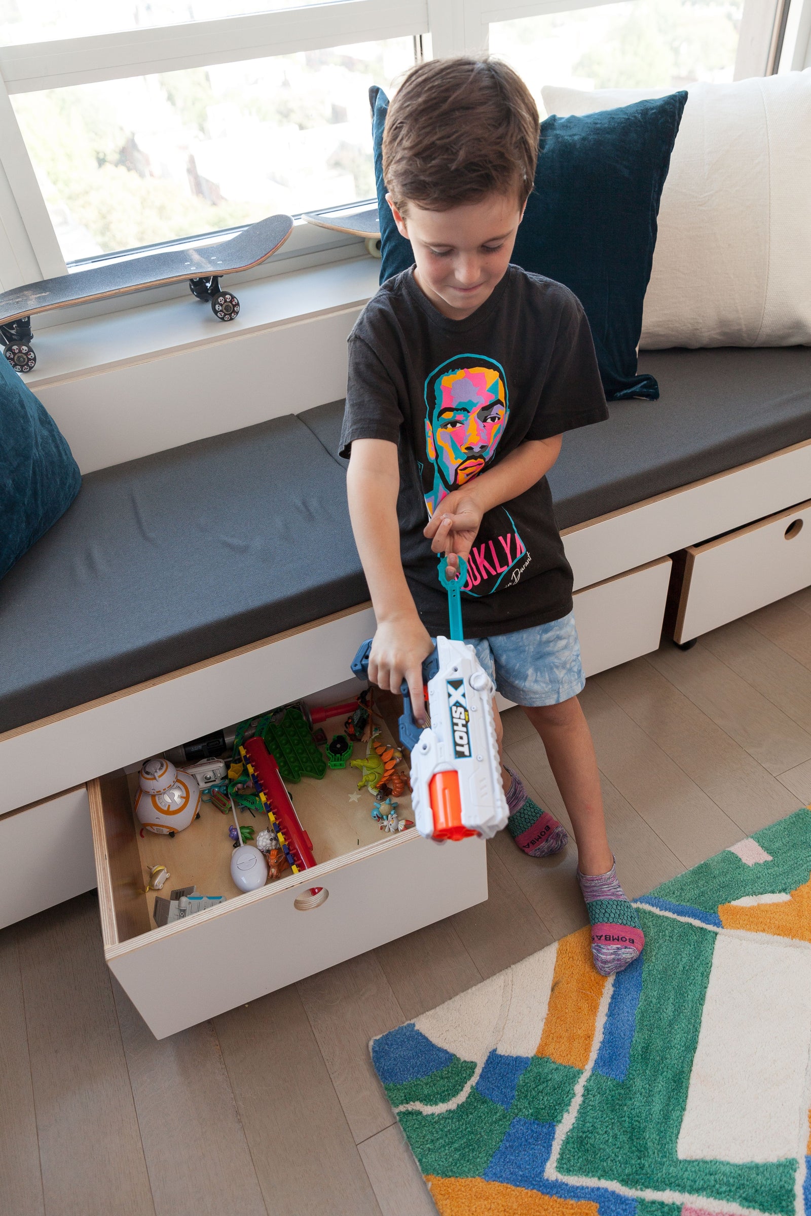 A cozy window seat with built-in storage features rolling toy carts for easy organization. A young boy plays with a toy blaster while an open drawer reveals a collection of action figures, LEGO pieces, and other toys. The cushioned seating area provides a functional and stylish solution for small spaces