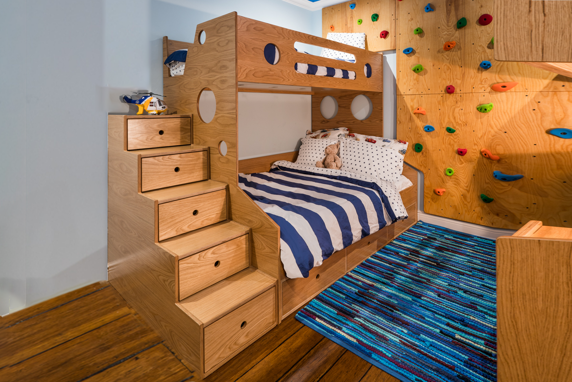 Marino Bunk Bed Twin over Full oak in wooden kid's room with custom rock climbing wall, blue bedding, white walls, and blue rug